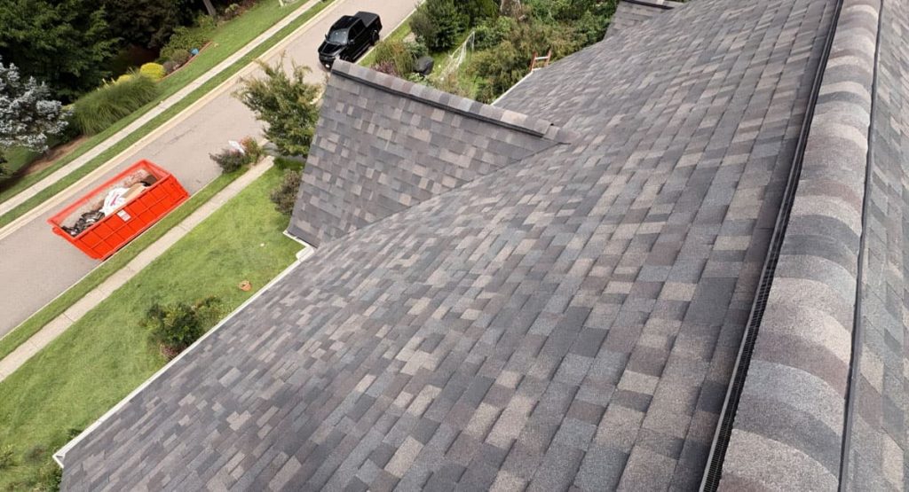 Aerial view of a gray shingle roof from above, with a red dumpster on the street and a parked black car nearby in a suburban neighborhood.