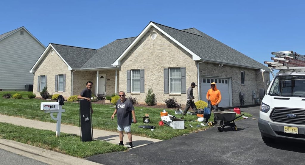 Group of workers cleaning up a front yard in front of a brick suburban house; tools, a wheelbarrow, and a ladder rest on a white service van parked in the driveway under a clear blue sky.