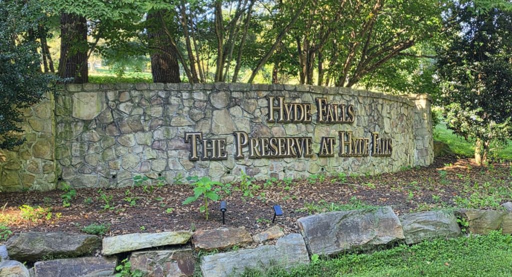 Stone exterior sign reading 'Hyde Falls The Preserve at Hyde Falls' set in a leafy park backdrop.